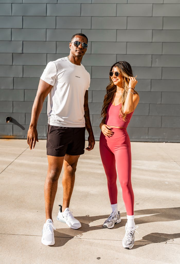 Two stylish individuals in sporty attire pose confidently outdoors in sunglasses.